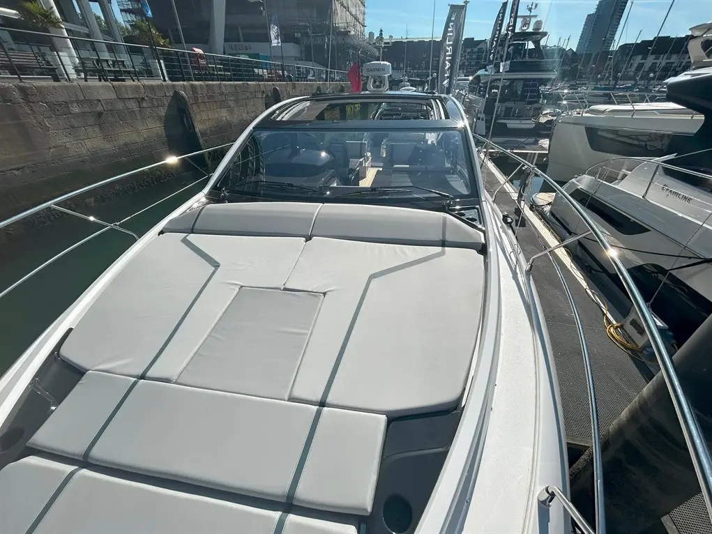 A sleek motorboat's deck with sunbathing cushions, showing a clear sky and a marina with other boats in the background.